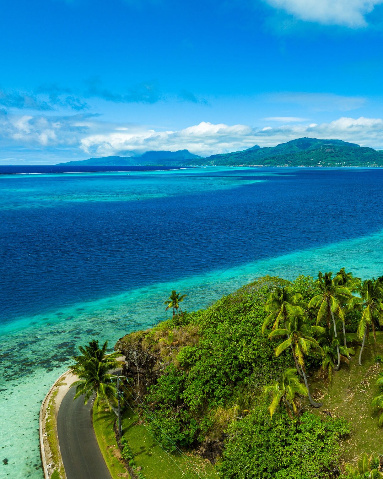 Aerial view of Taha’a in French Polynesia, showing a coastal road lined with palm trees, turquoise lagoon and distant green mountains.