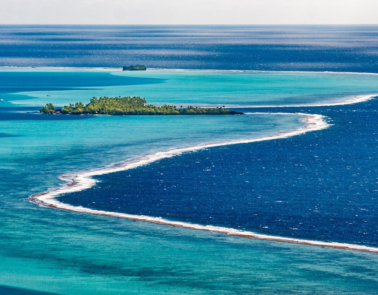 Aerial view of Taha’a’s barrier reef curving through turquoise and deep blue waters with small palm-covered islets scattered beyond.