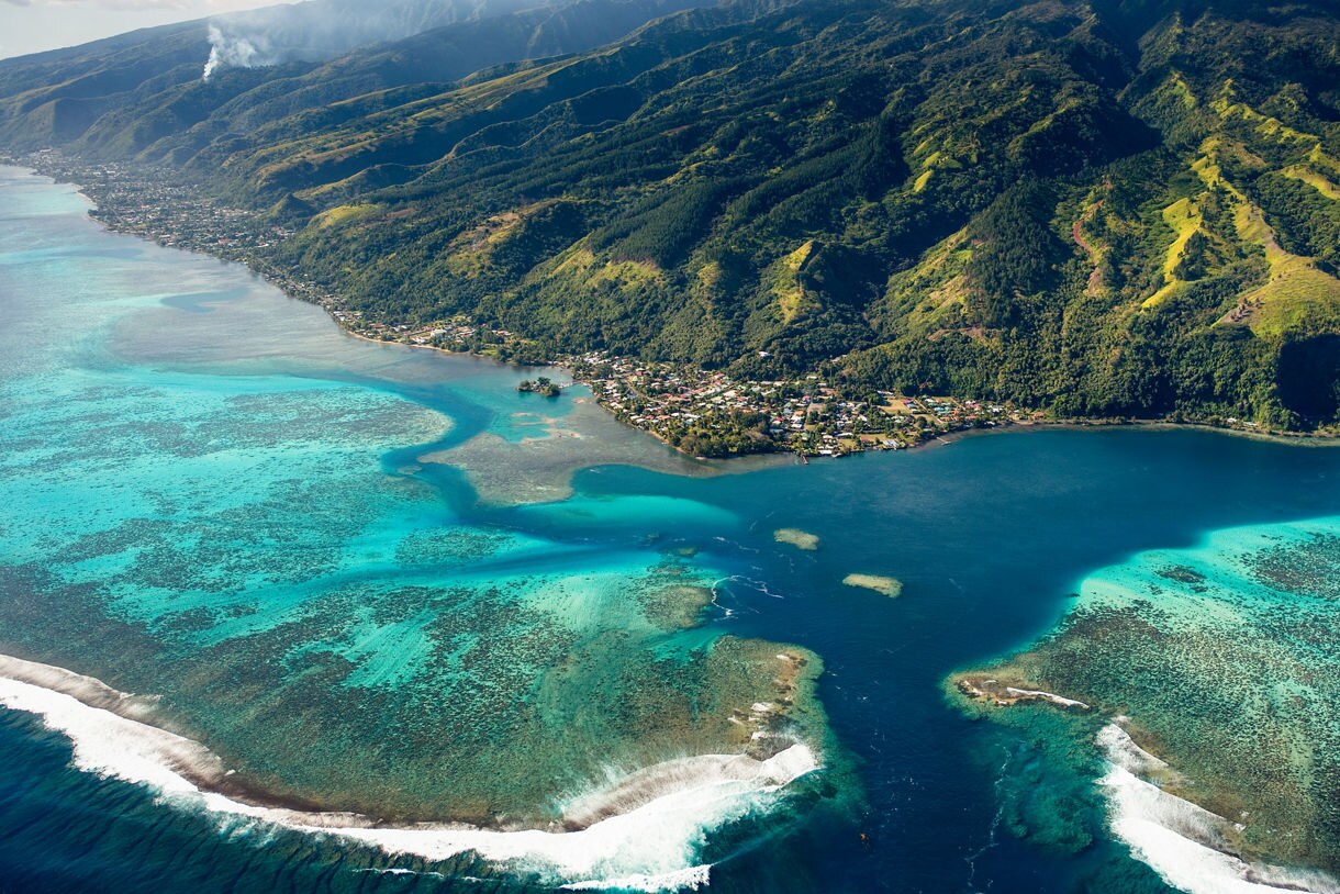 Aerial view of Tahiti’s coastline with bright turquoise shallows, coral reefs, deep blue lagoon water and steep green mountains dotted with small coastal villages.