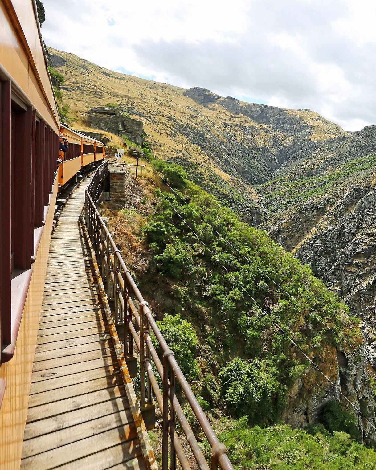 A yellow train curving along a wooden bridge through Taieri Gorge, surrounded by rugged cliffs and a narrow river below.