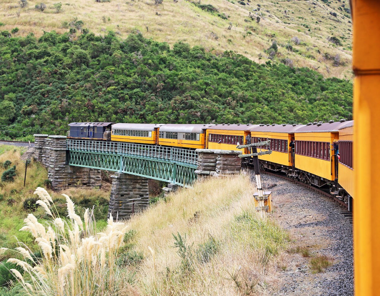 Yellow passenger train crossing a green iron bridge through the steep, grassy hills of Taieri Gorge.