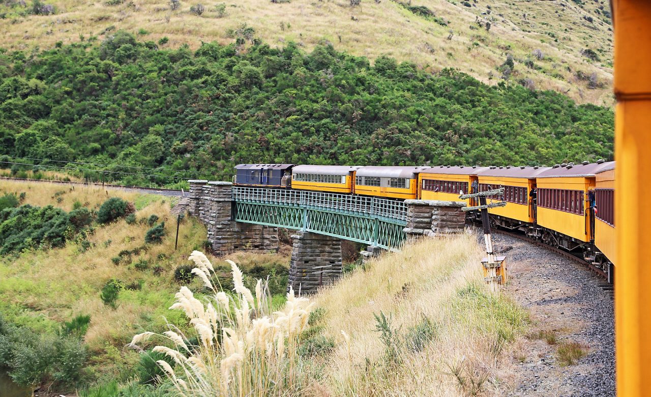 Yellow passenger train crossing a green iron bridge through the steep, grassy hills of Taieri Gorge.