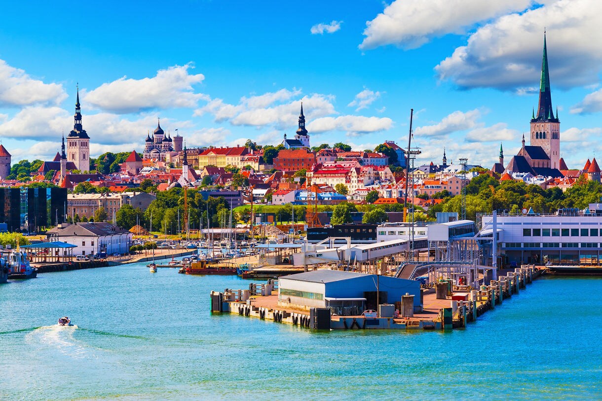 Panoramic view of Tallinn, Estonia, showing the harbor with boats in the foreground and the Old Town skyline with church spires, pastel buildings and red rooftops under a bright blue sky.