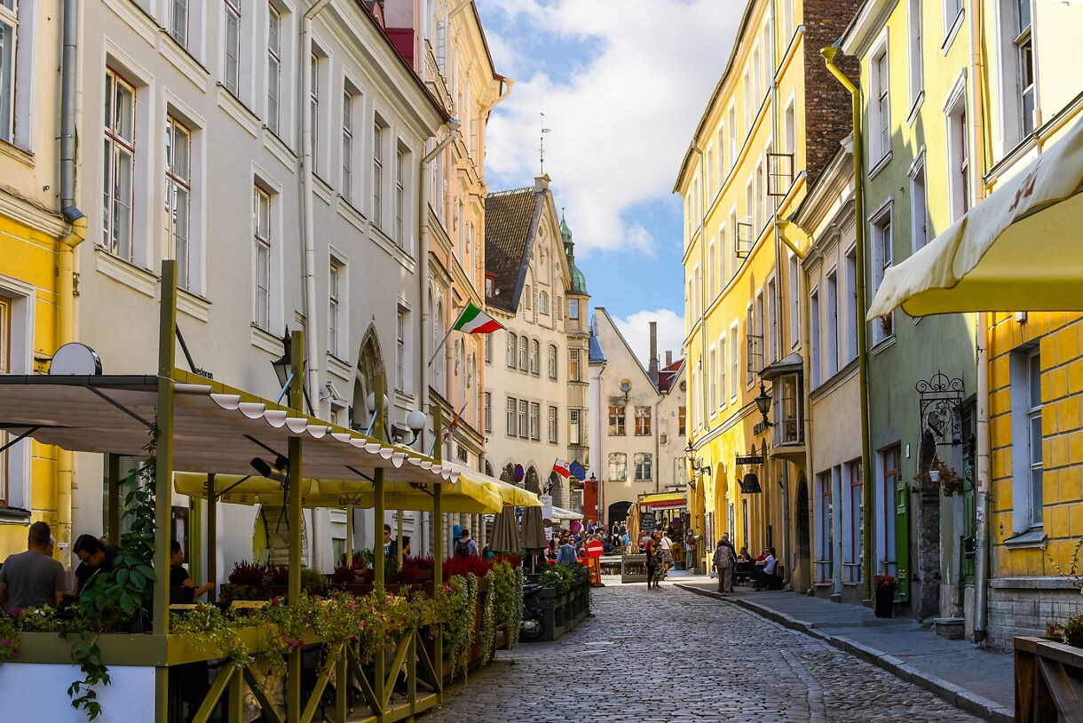 Narrow cobblestone street in Tallinn, Estonia, with pastel-colored historic buildings, outdoor café seating and pedestrians enjoying the lively Old Town atmosphere.