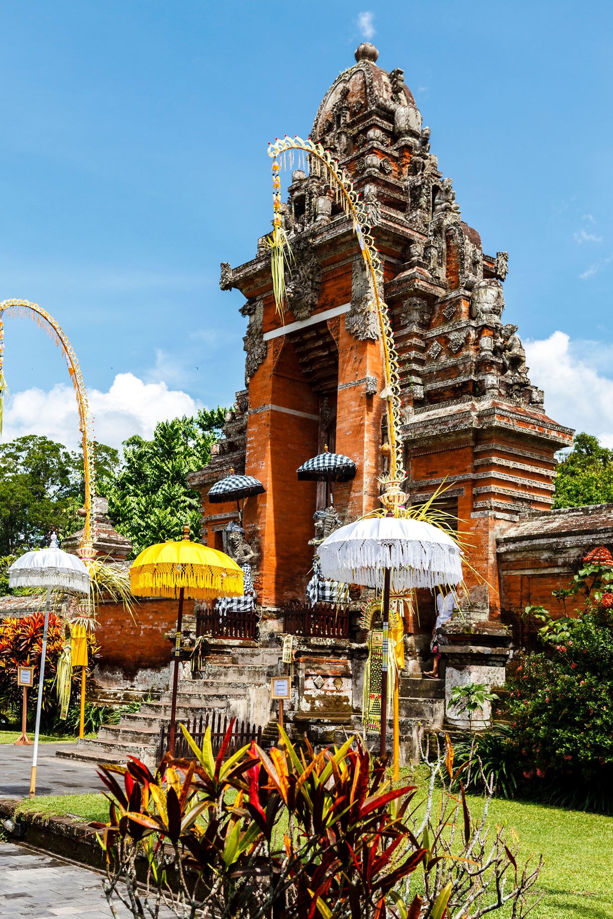 Traditional Balinese temple entrance built of orange brick and detailed stone carvings, surrounded by yellow and white ceremonial umbrellas, tall decorative penjor poles and lush garden greenery beneath a clear blue sky.