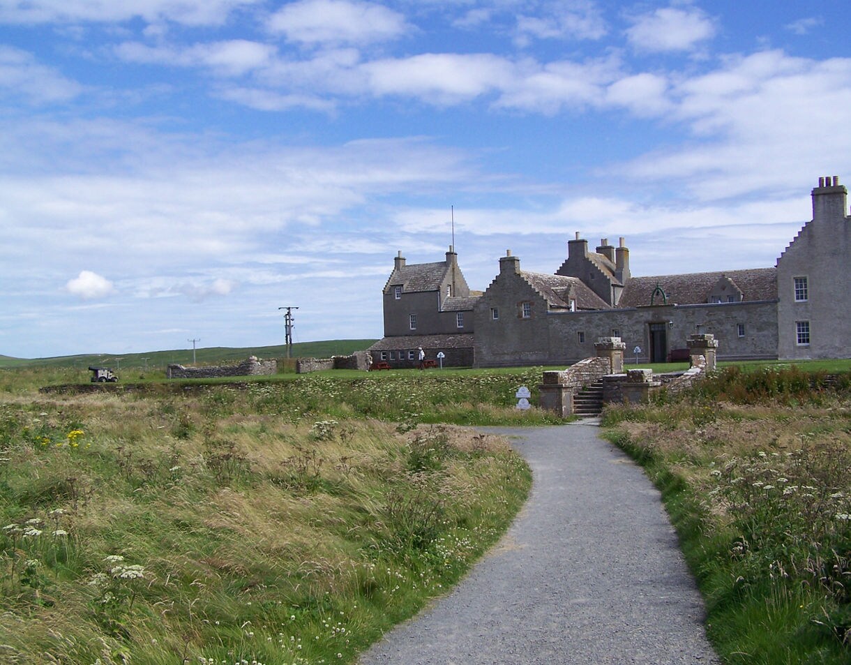 Tankerness House Museum in Kirkwall, a historic stone building with gabled roofs set against open grassy fields and a partly cloudy sky.