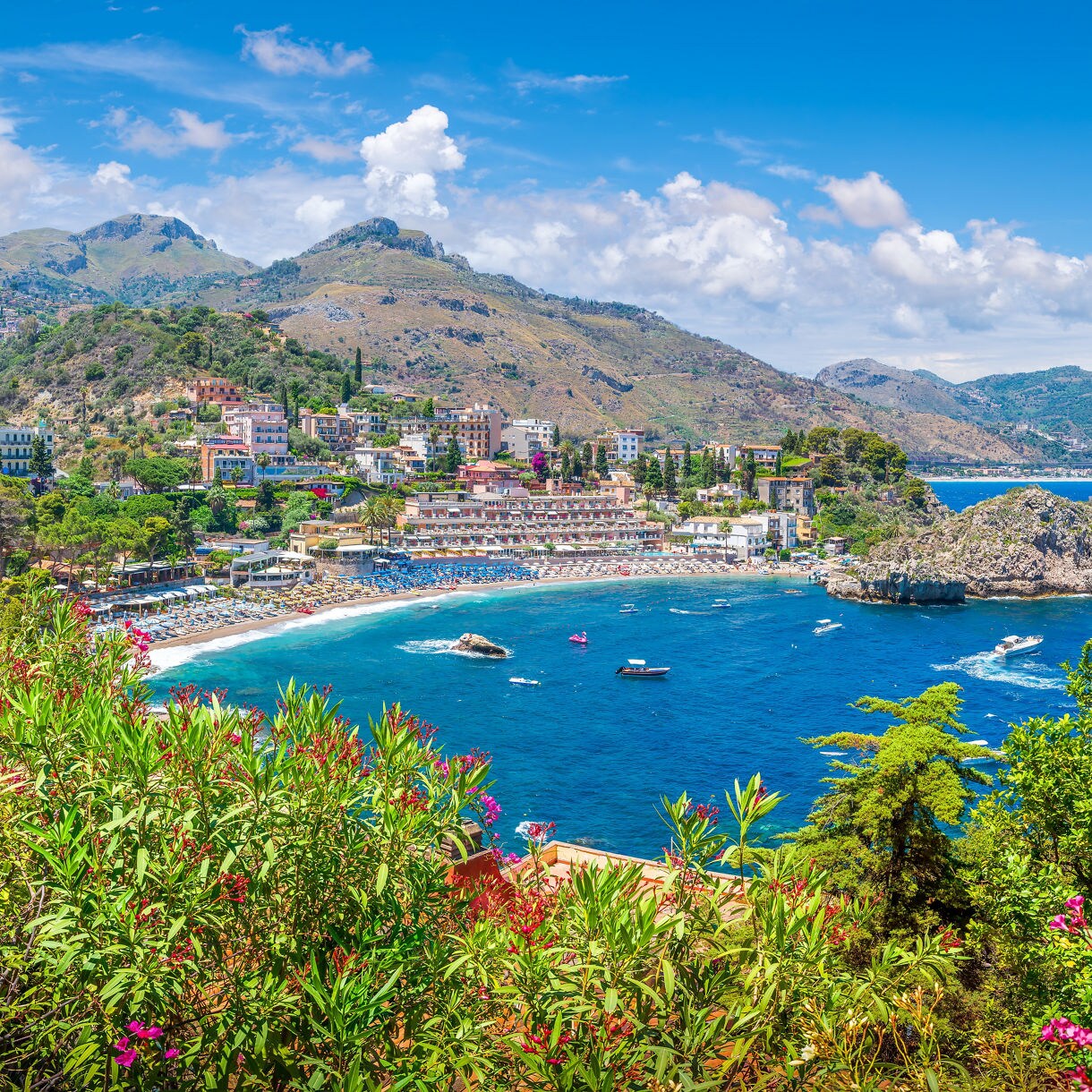 Panoramic view of Taormina’s curved beach and rocky islet surrounded by bright blue water, framed by lush greenery and pink flowers with rugged hills rising in the background.