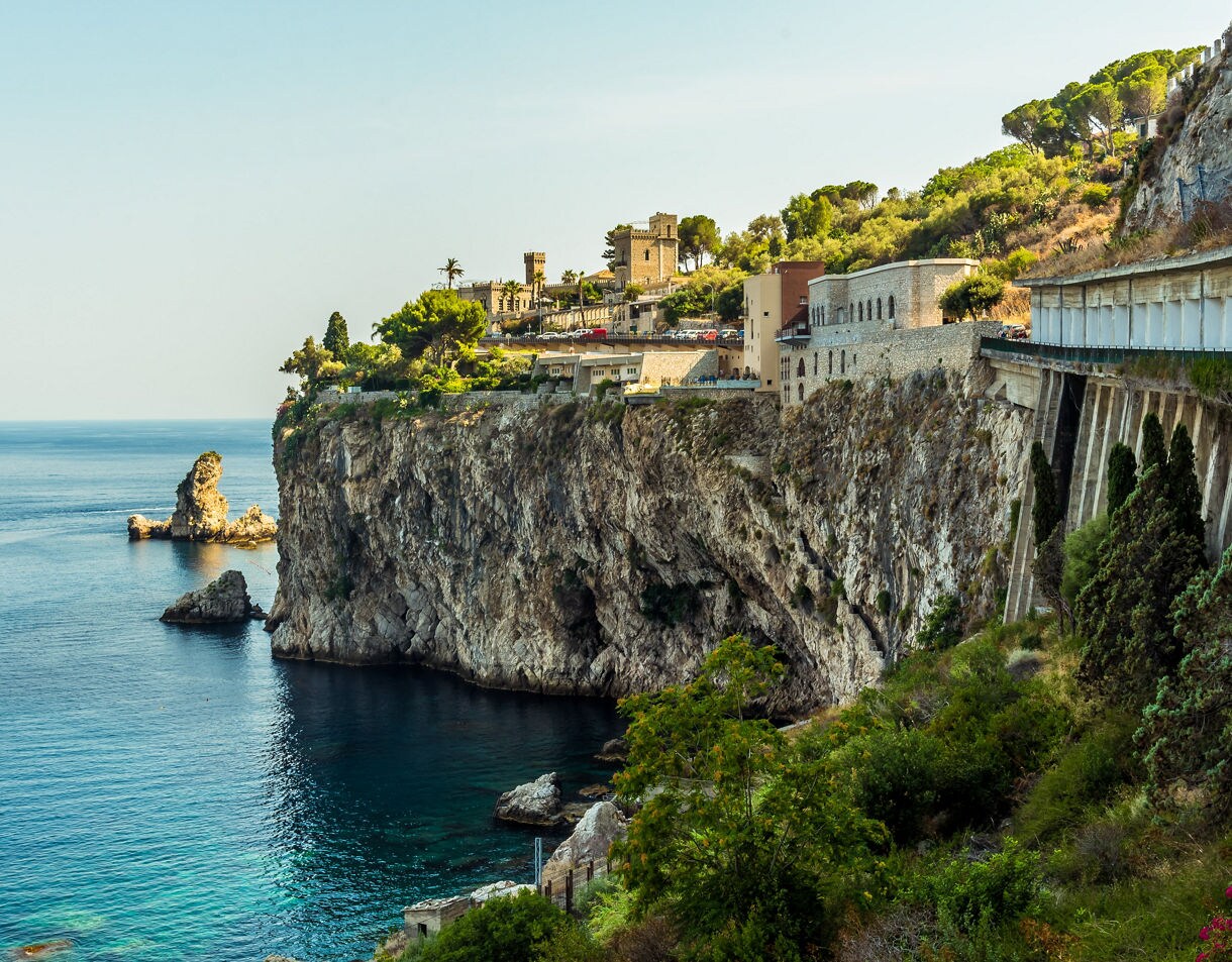 Steep rocky cliffs overlooking calm turquoise water near Taormina, with historic stone buildings and greenery along the cliff edge.