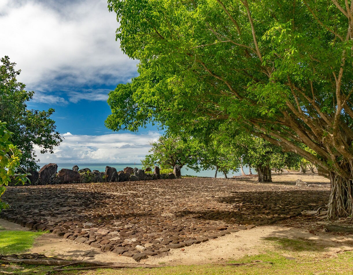 Stone courtyard of Taputapuātea Marae in Raiatea, surrounded by large trees and overlooking the blue South Pacific.