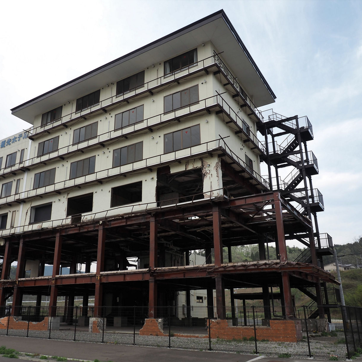 Weathered multi-story concrete hotel in Taro with its lower floors stripped to a steel frame after tsunami damage, standing fenced off against a cloudy sky.