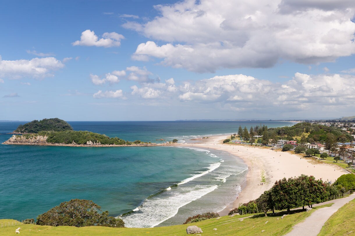 Panoramic view from Mount Maunganui overlooking a curved sandy beach, the blue ocean, a small island and the city of Tauranga with buildings and a harbor in the distance.