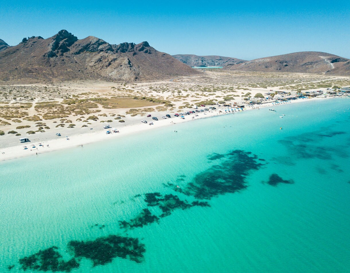 Aerial view of Tecolote Beach with clear turquoise water, sandy shoreline, parked cars and rugged desert mountains in the background.