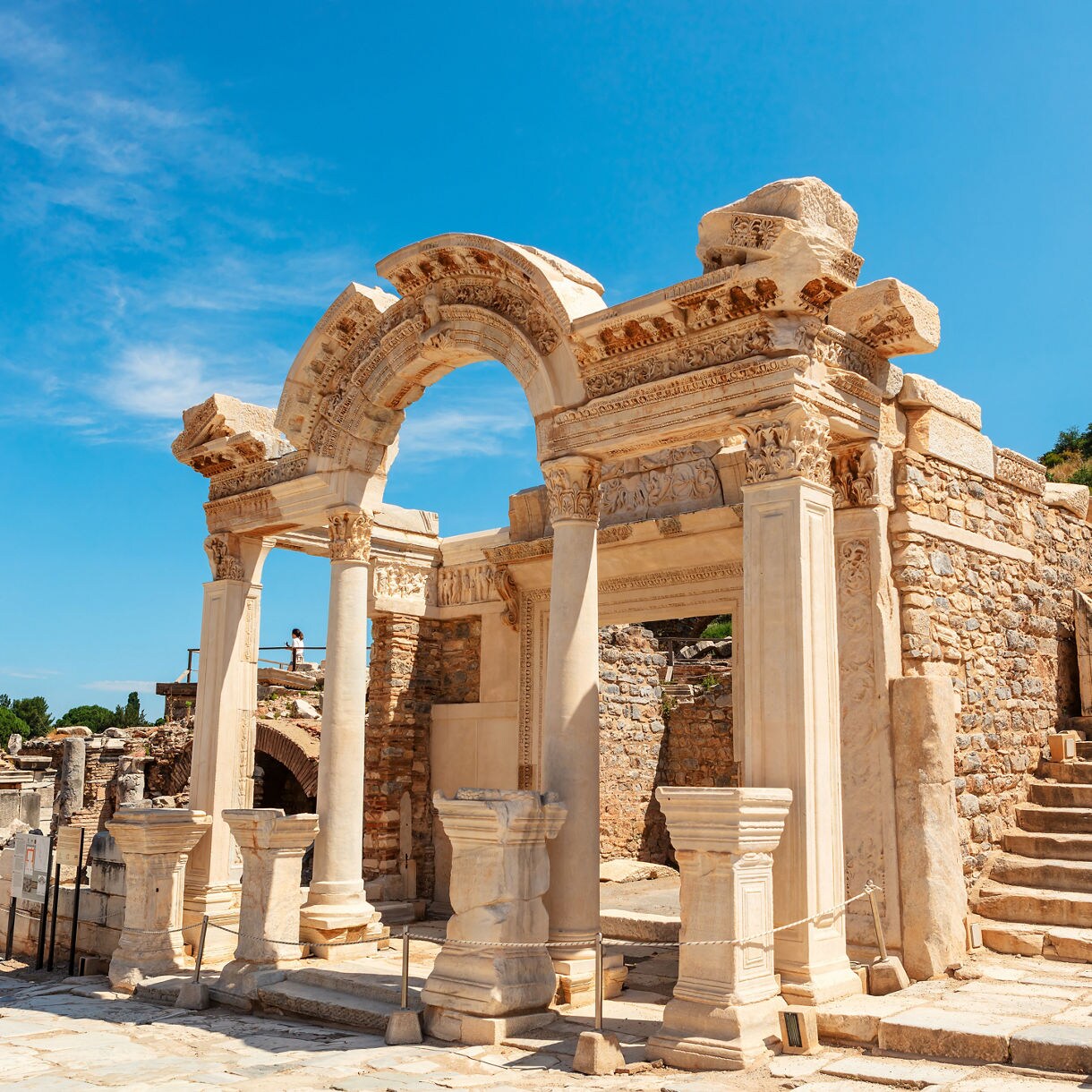 Ancient marble archway and columns of the Temple of Hadrian in Ephesus, Turkey, surrounded by sunlit stone ruins under a bright blue sky.