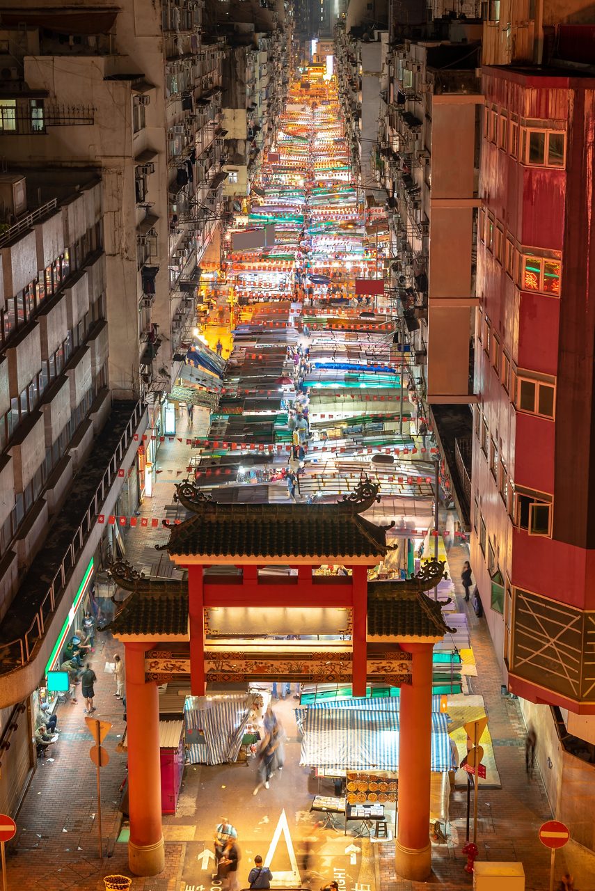 Aerial view of Temple Street Night Market in Hong Kong at night, with a traditional red gate and rows of colorful illuminated stalls between tall buildings.