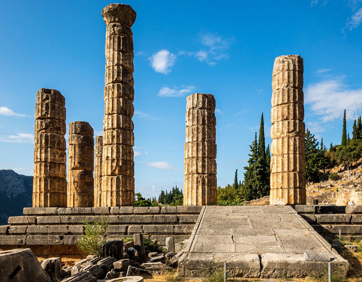 Ruins of the Temple of Apollo in Delphi, Greece, featuring tall stone columns against a backdrop of trees and mountains.