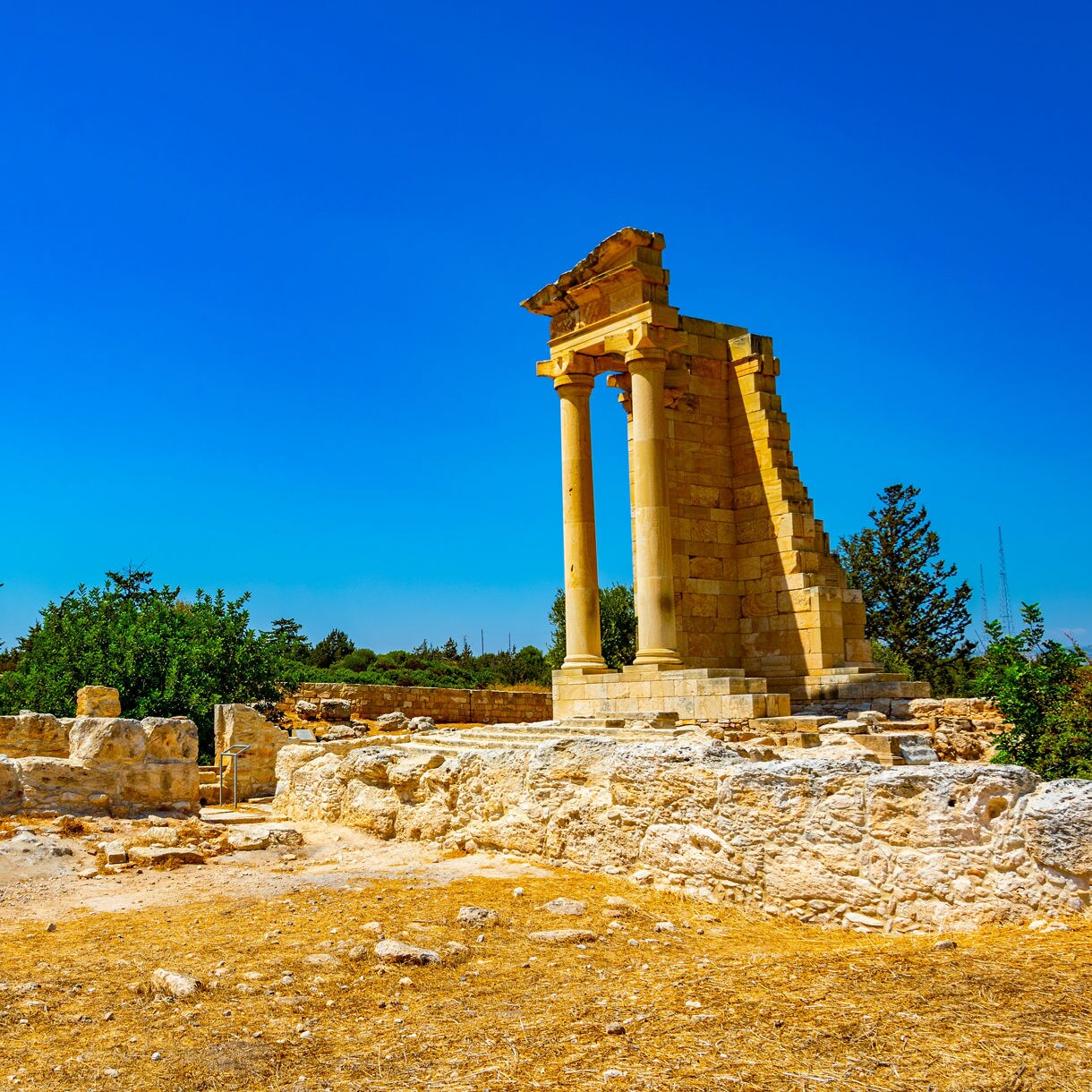Ruins of the ancient Temple of Apollo in Cyprus, featuring a partially standing portico with tall stone columns surrounded by low excavated walls, dry golden earth and scattered shrubs under a bright blue sky.