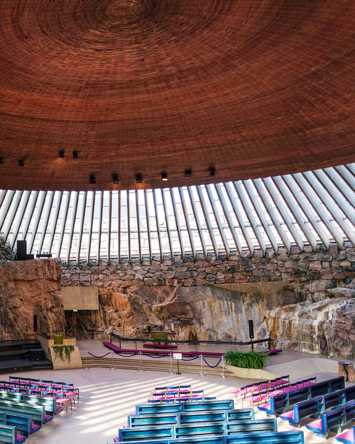 Interior of Temppeliaukio Church in Helsinki, featuring rugged rock walls, a circular copper dome ceiling and rows of blue pews surrounding the altar.