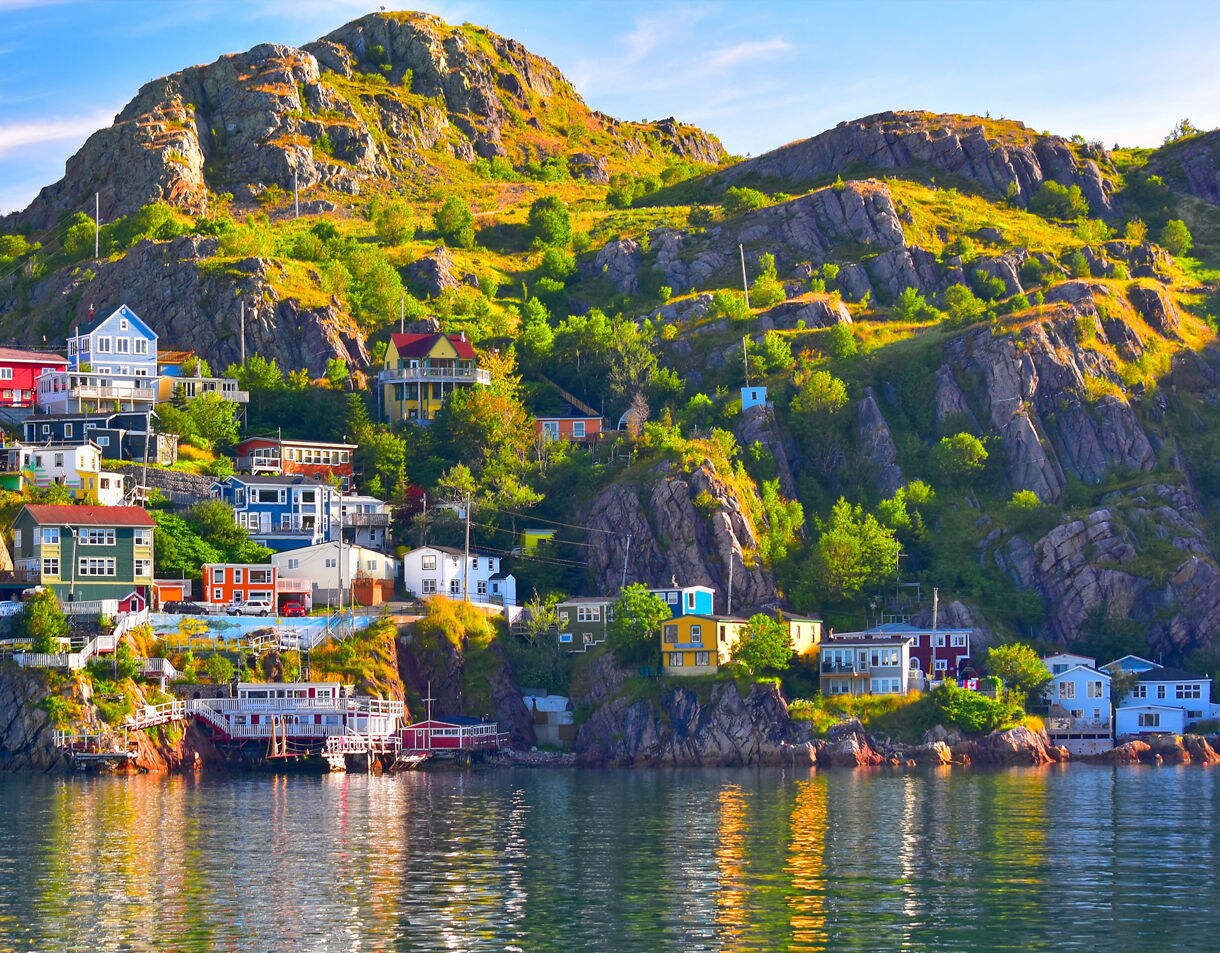 A hillside of colorful houses set along a rocky shoreline in St. John’s, Newfoundland, with vivid greens and warm sunlight reflecting across calm harbor water.