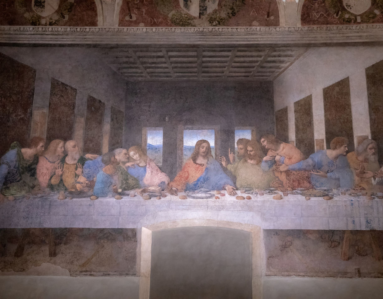 Mural of The Last Supper by Leonardo da Vinci depicting Jesus and his disciples seated at a long table in a dimly lit room with open windows in the background.