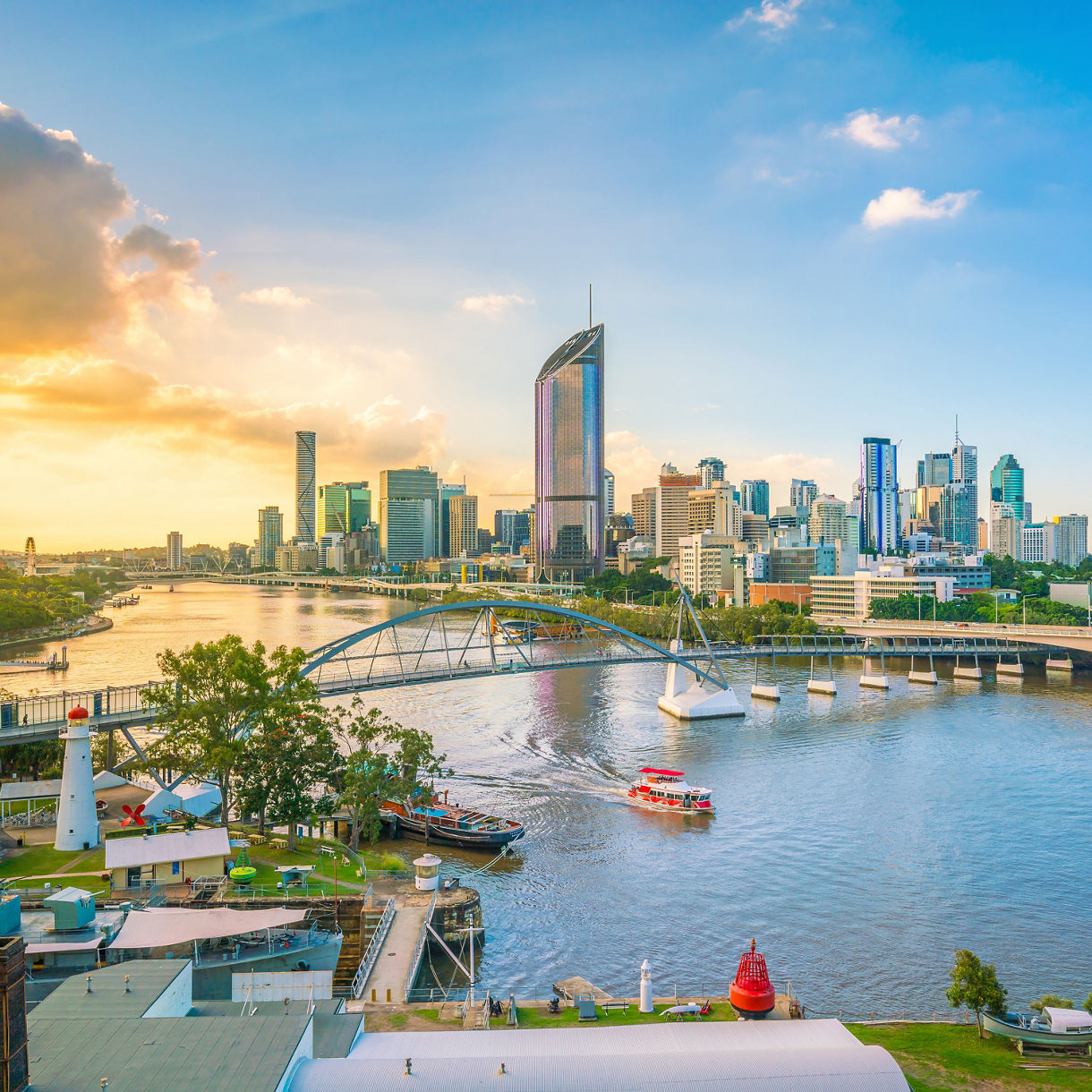 Set dining tables under black umbrellas on an outdoor riverfront patio, with the blurred steel frame of Brisbane’s Story Bridge spanning across the water in the background.