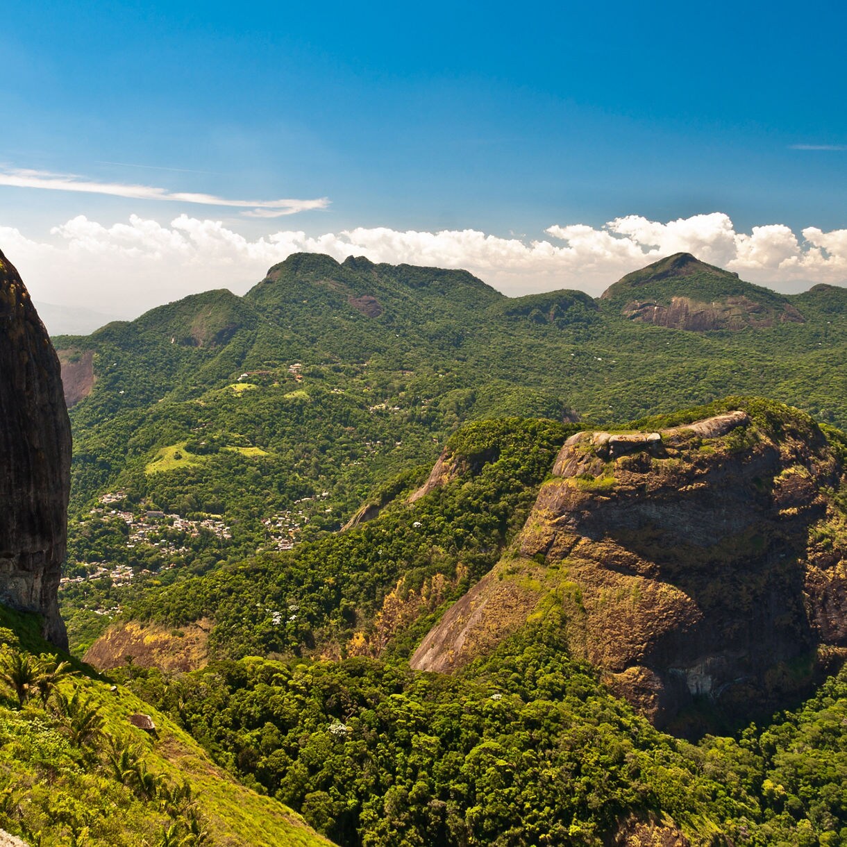 Expansive view of Tijuca Forest with dense green hills, steep rocky cliffs and a bright blue sky overhead.