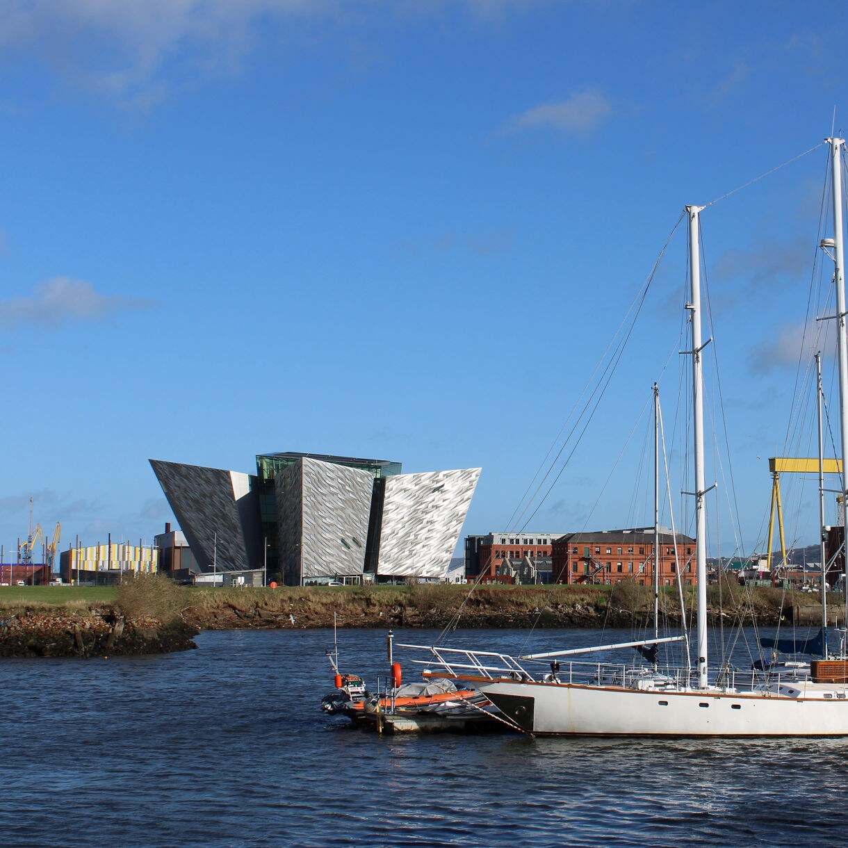 White sailboat docked in front of the modern angular Titanic Belfast museum with Harland & Wolff shipyard crane in the background.