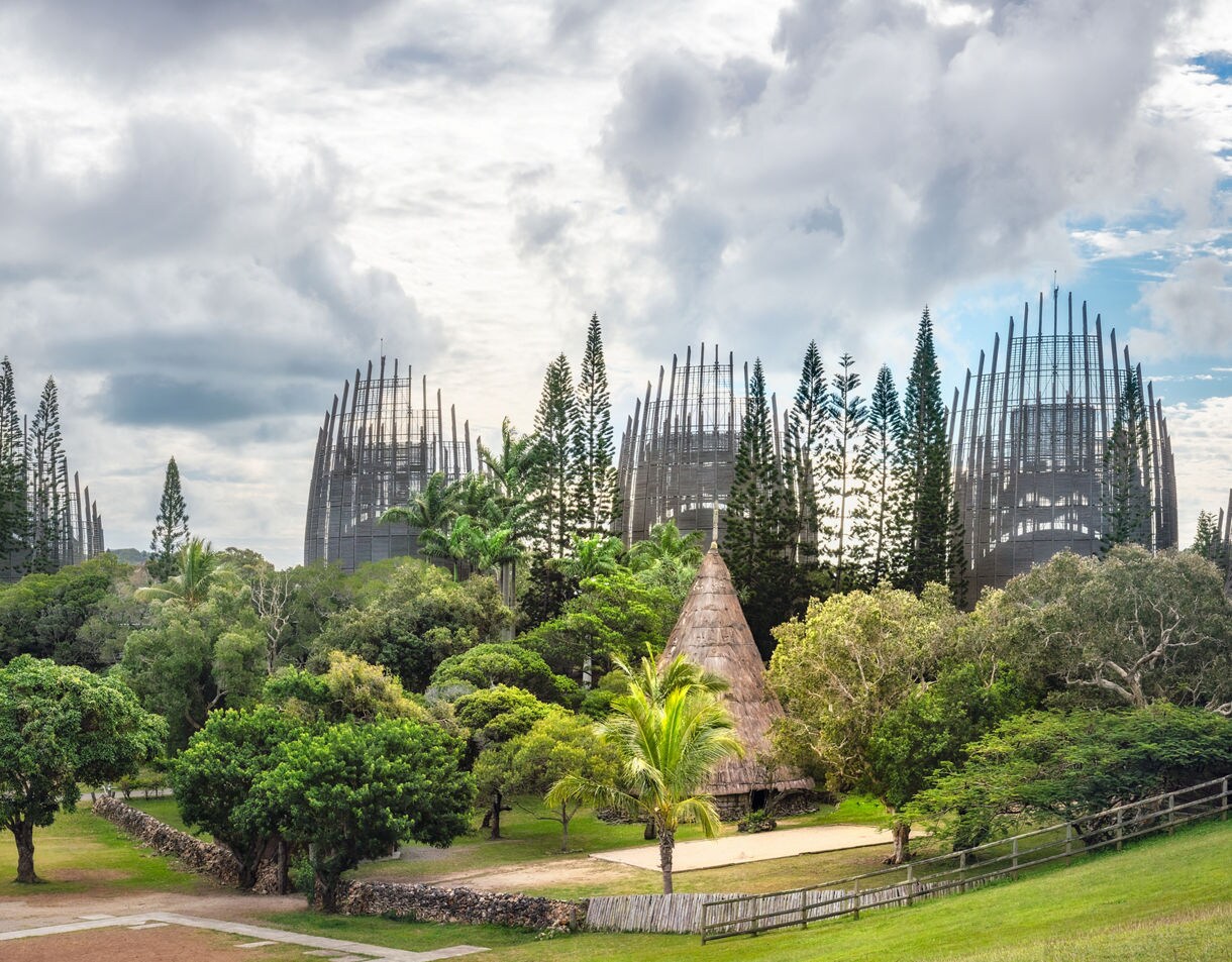 Panoramic view of the Tjibaou Cultural Centre, featuring tall wooden pavilions shaped like contemporary Kanak huts surrounded by dense green trees and an open grassy landscape under a cloudy sky.