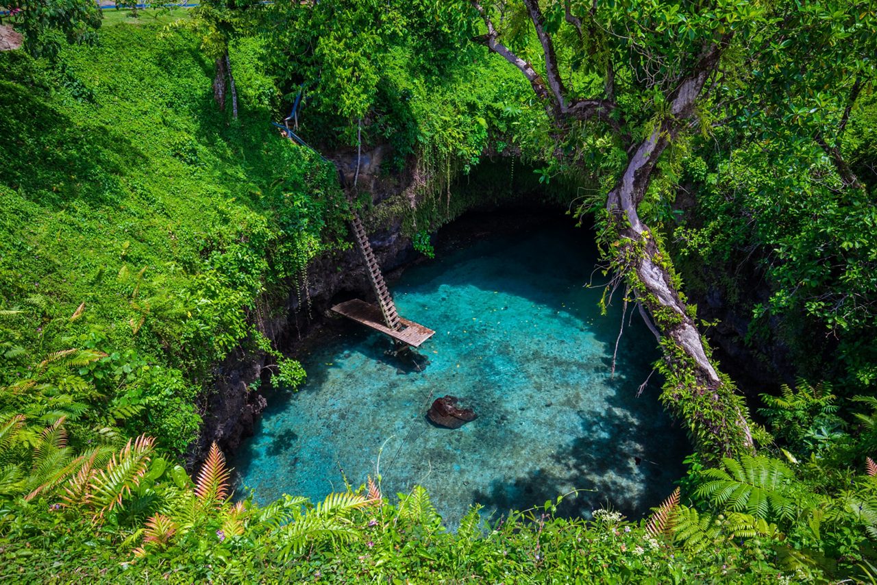 A deep turquoise swimming hole in Samoa with a wooden ladder and platform, encircled by lush green vegetation.
