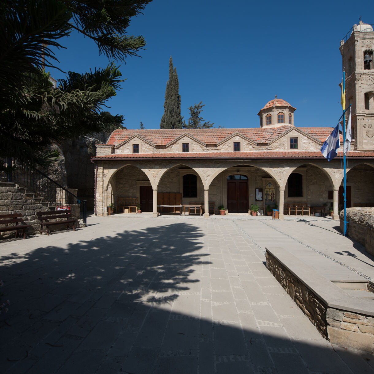 Stone monastery in Tochni, Cyprus, featuring a long arched portico, a central wooden doorway, terracotta roof tiles and a tall bell tower under a clear blue sky.