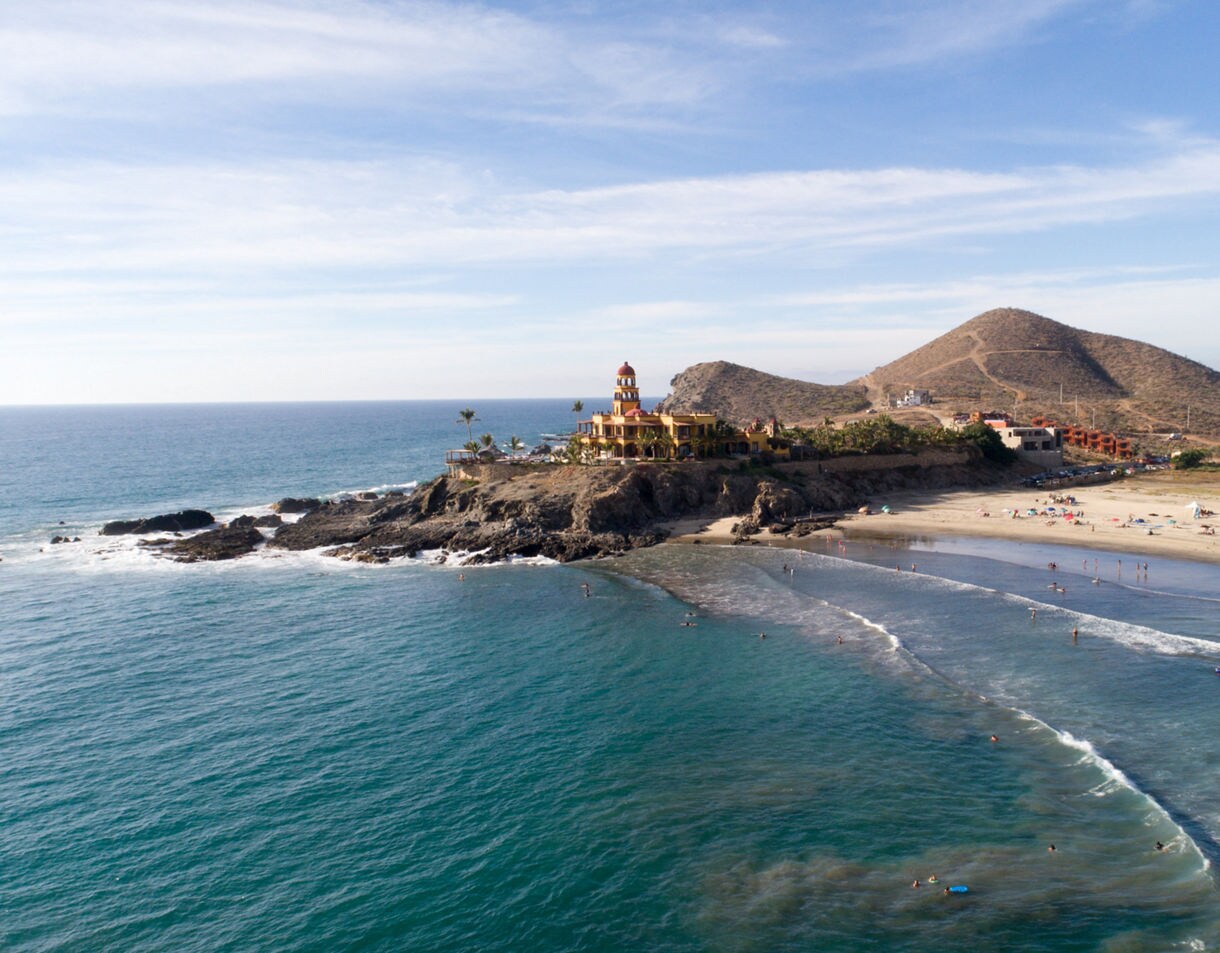 Aerial view of Todos Santos beach with turquoise waves, sandy shore and a colorful coastal building set against desert hills.