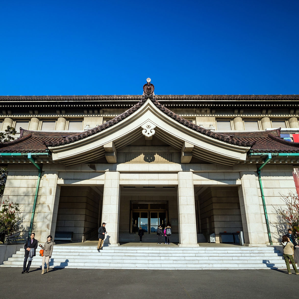 Front facade of the Tokyo National Museum, featuring a curved traditional Japanese roof atop a stone building, with visitors on the entrance steps under a clear blue sky.