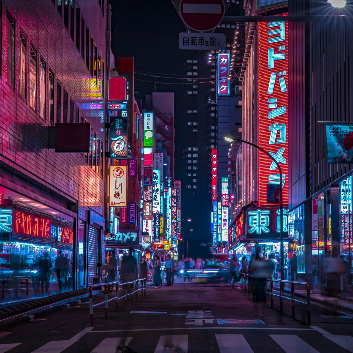 A busy Tokyo street at night lined with glowing neon signs in Japanese, with pedestrians crossing below towering illuminated buildings