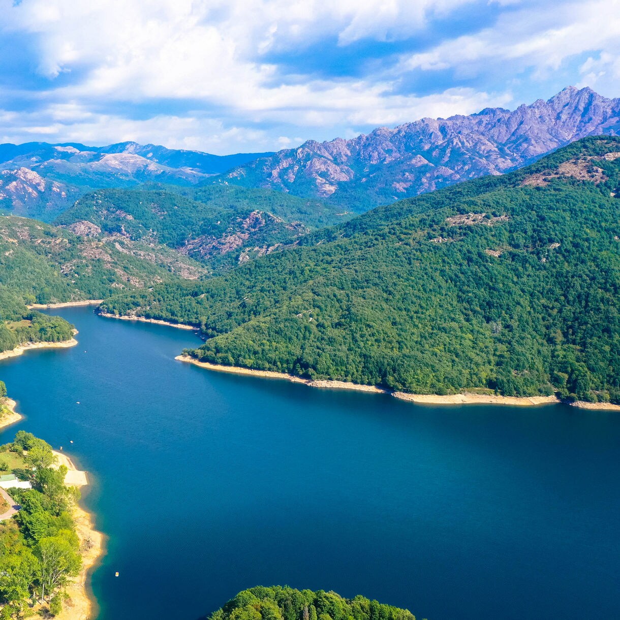 Aerial view of Lake Tolla in Corsica, framed by dense green hills, a small village and rugged mountain peaks under a bright sky.