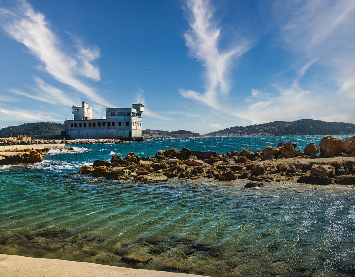Rocky shoreline with turquoise water rippling under bright sun, an old white coastal building on a breakwater and forested hills rising across the bay.