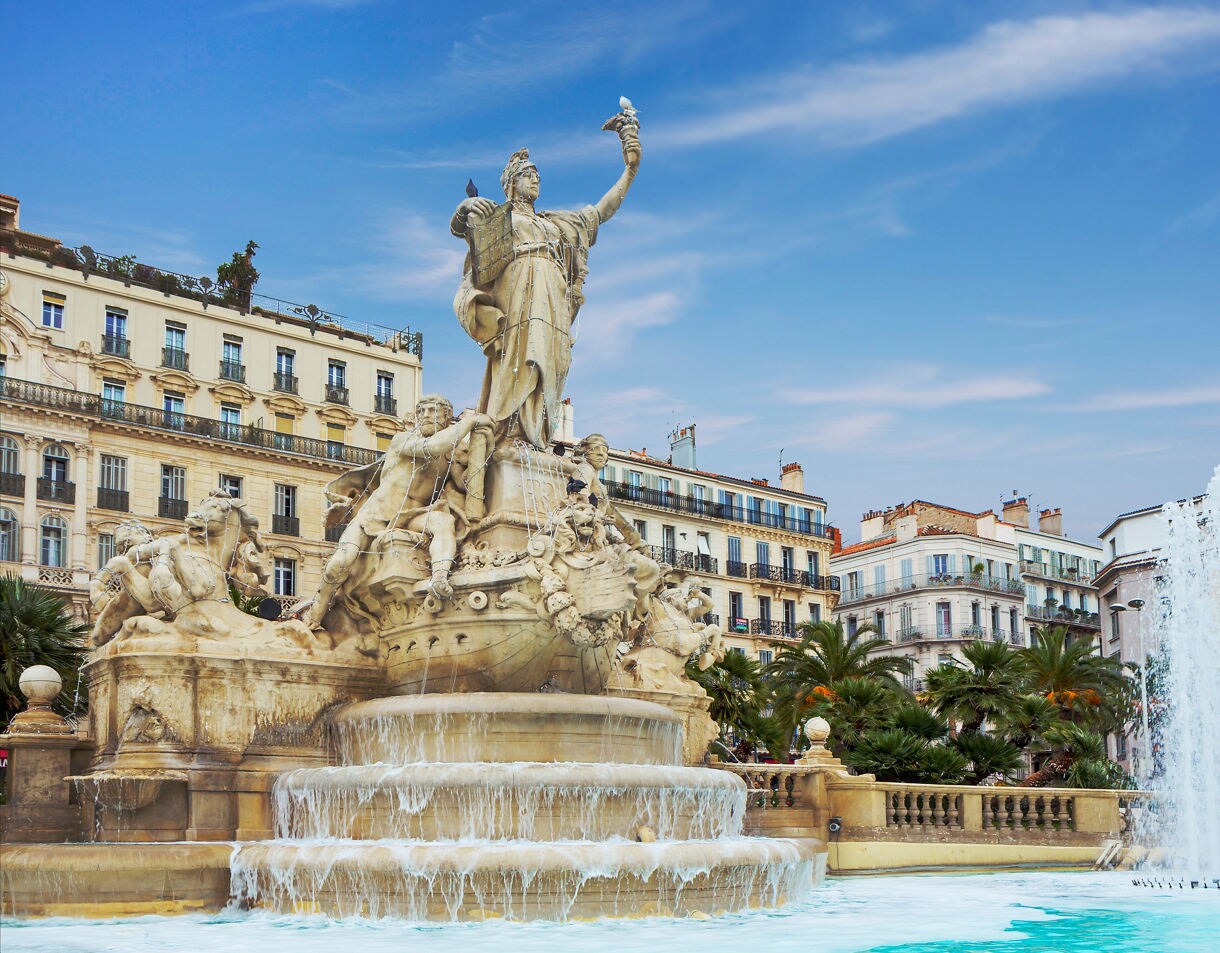 Elaborate marble fountain in Toulon featuring mythological figures, flowing water and a central female statue holding a bird, set against classic French buildings and a blue sky.