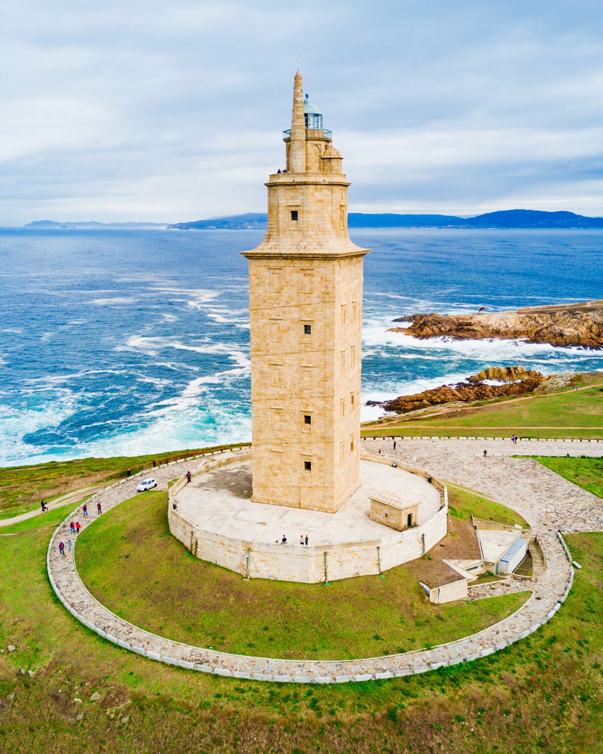 Aerial view of the Tower of Hercules in A Coruña, Spain, a tall stone lighthouse on a grassy headland overlooking rocky coastline and blue ocean waves.