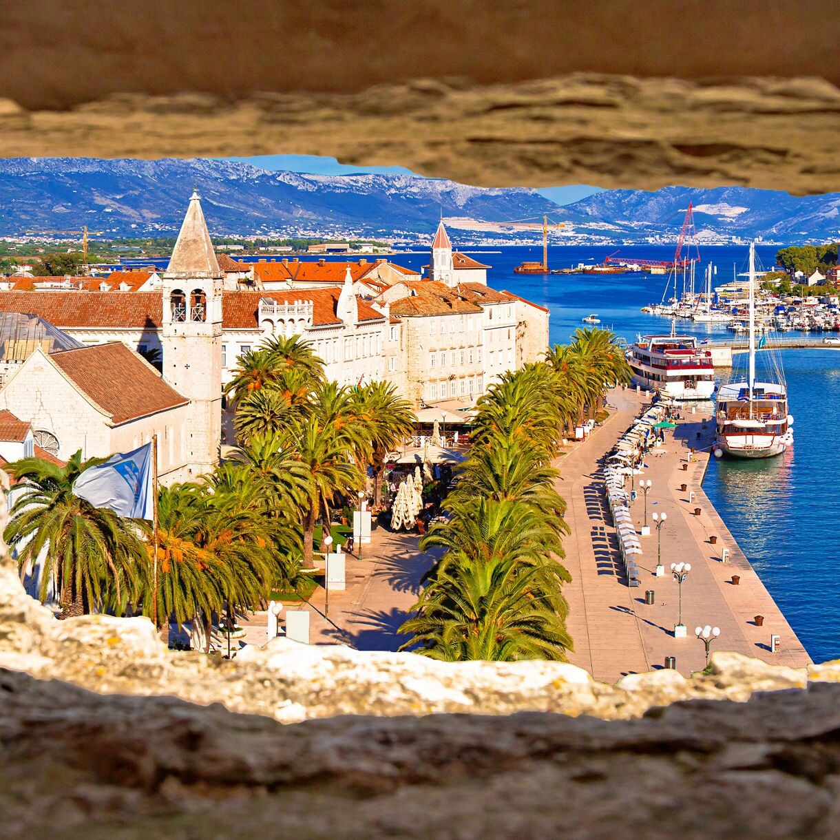 View through a stone window opening of Trogir’s waterfront, showing palm trees, historic buildings with red-tiled roofs, boats docked along the promenade and mountains in the distance.