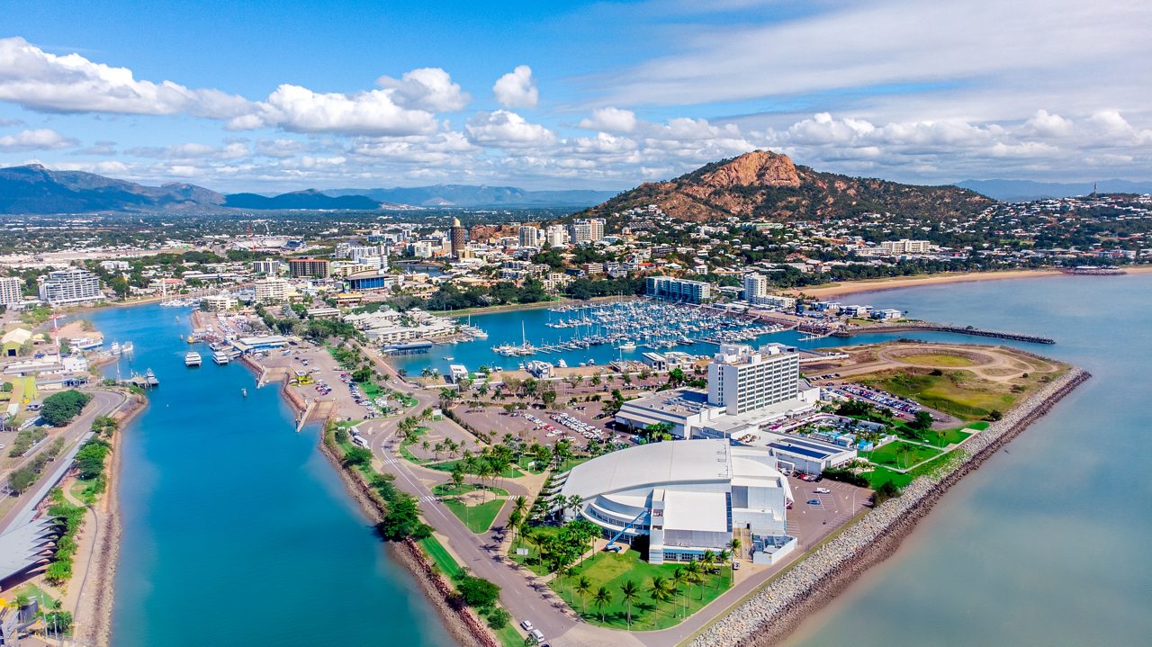 A bright aerial view of Townsville, Australia, showing a marina filled with boats, a curved shoreline and the red cliffs of Castle Hill rising behind the city under a blue sky.
