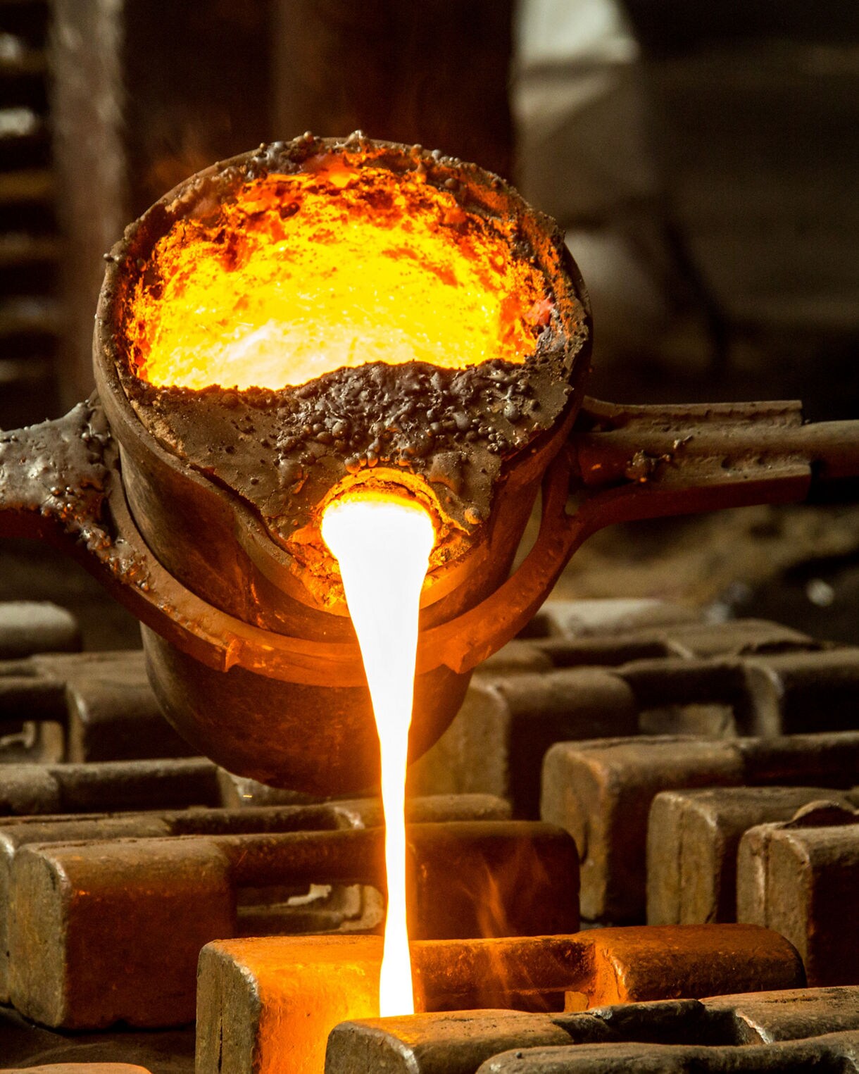 A glowing crucible tilting to release a stream of hot molten metal into waiting molds, with sparks and warm orange light illuminating the workshop.