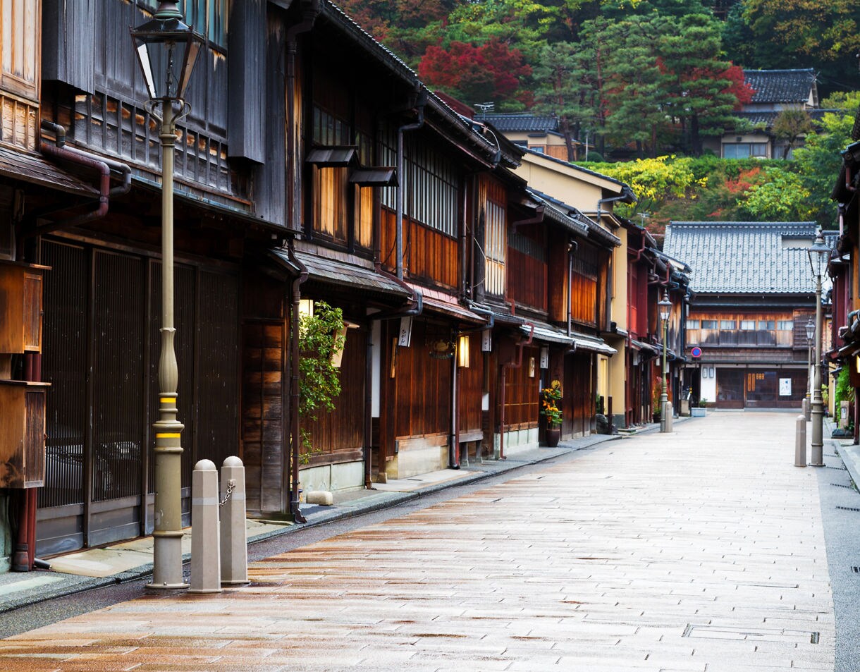 A quiet street bordered by traditional wooden buildings with sliding doors and lanterns, set beneath colorful hillside trees after a light rain.