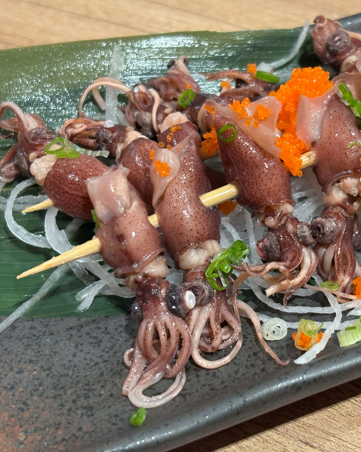 A plate of small whole squid skewered and arranged over shredded daikon with bright orange roe, sliced green onions and a lemon wedge on a dark platter.