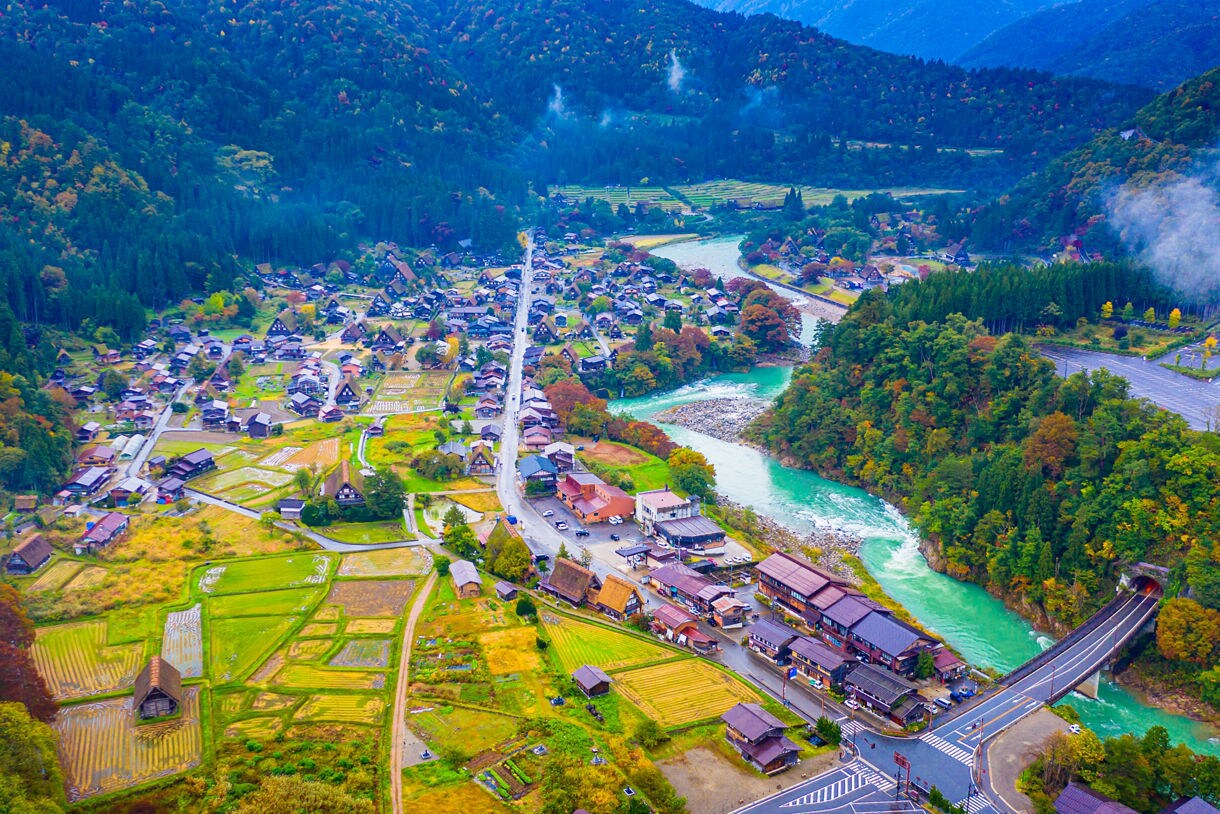 A wide aerial view of a traditional Japanese village with steep thatched houses, colorful fields and a vivid blue-green river surrounded by dense mountain forests.