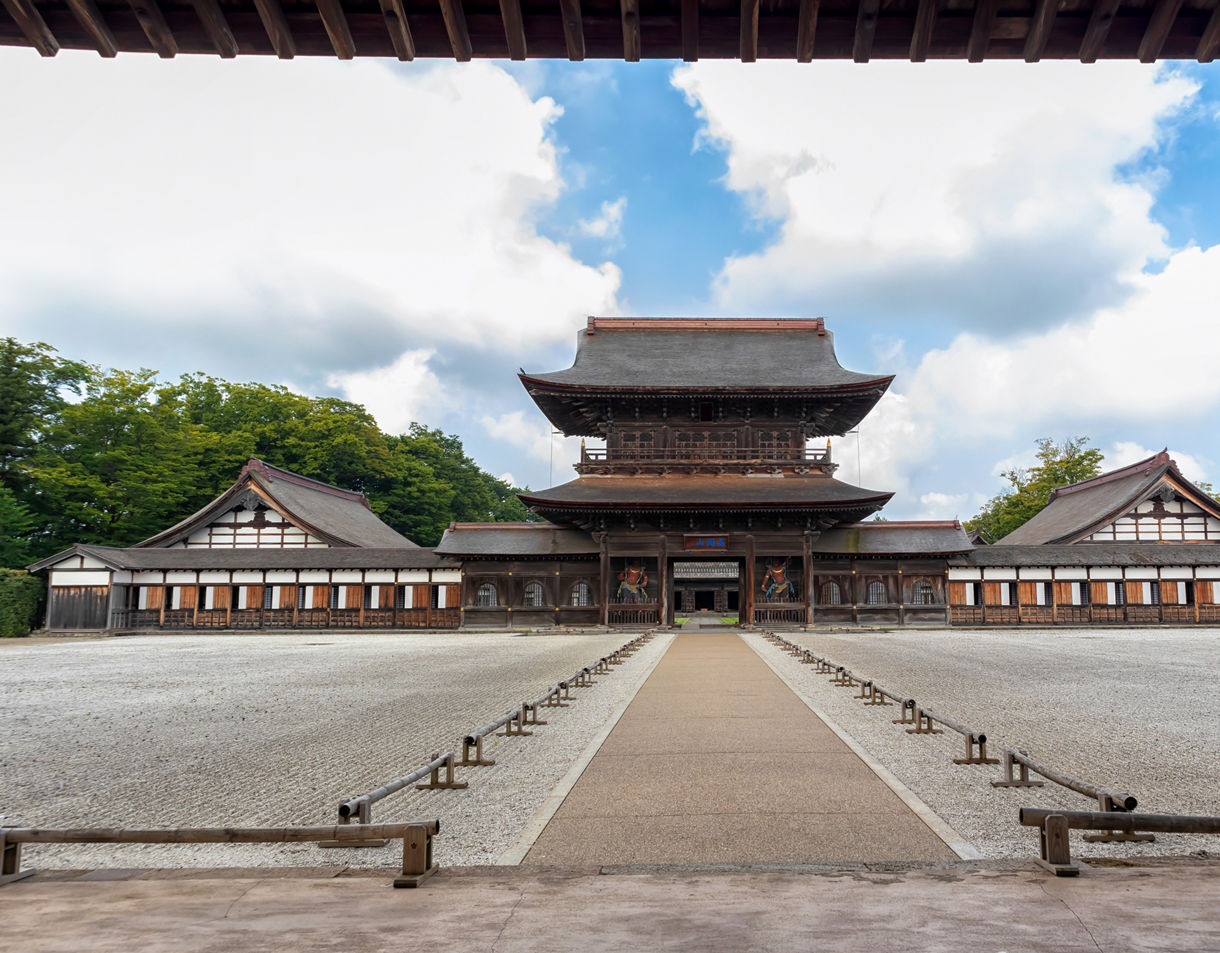 A large wooden temple with sweeping tiered roofs centered at the end of a straight gravel walkway, flanked by long side halls and framed by trees under a bright sky.