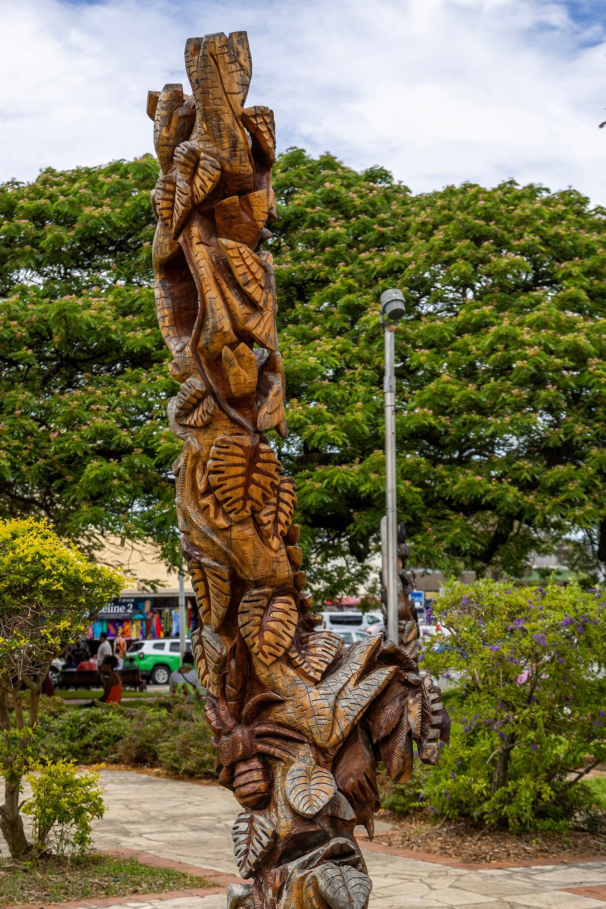 Close-up of a tall wooden totem covered in detailed leaf and face carvings, standing on a grassy patch with trees and a walkway in the background.