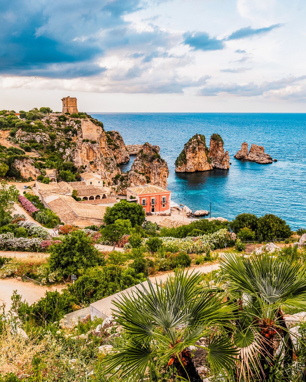 View of a coastal village in Sicily with rocky sea stacks rising from turquoise water, a small cluster of historic buildings, lush greenery and a lookout tower on a cliff.
