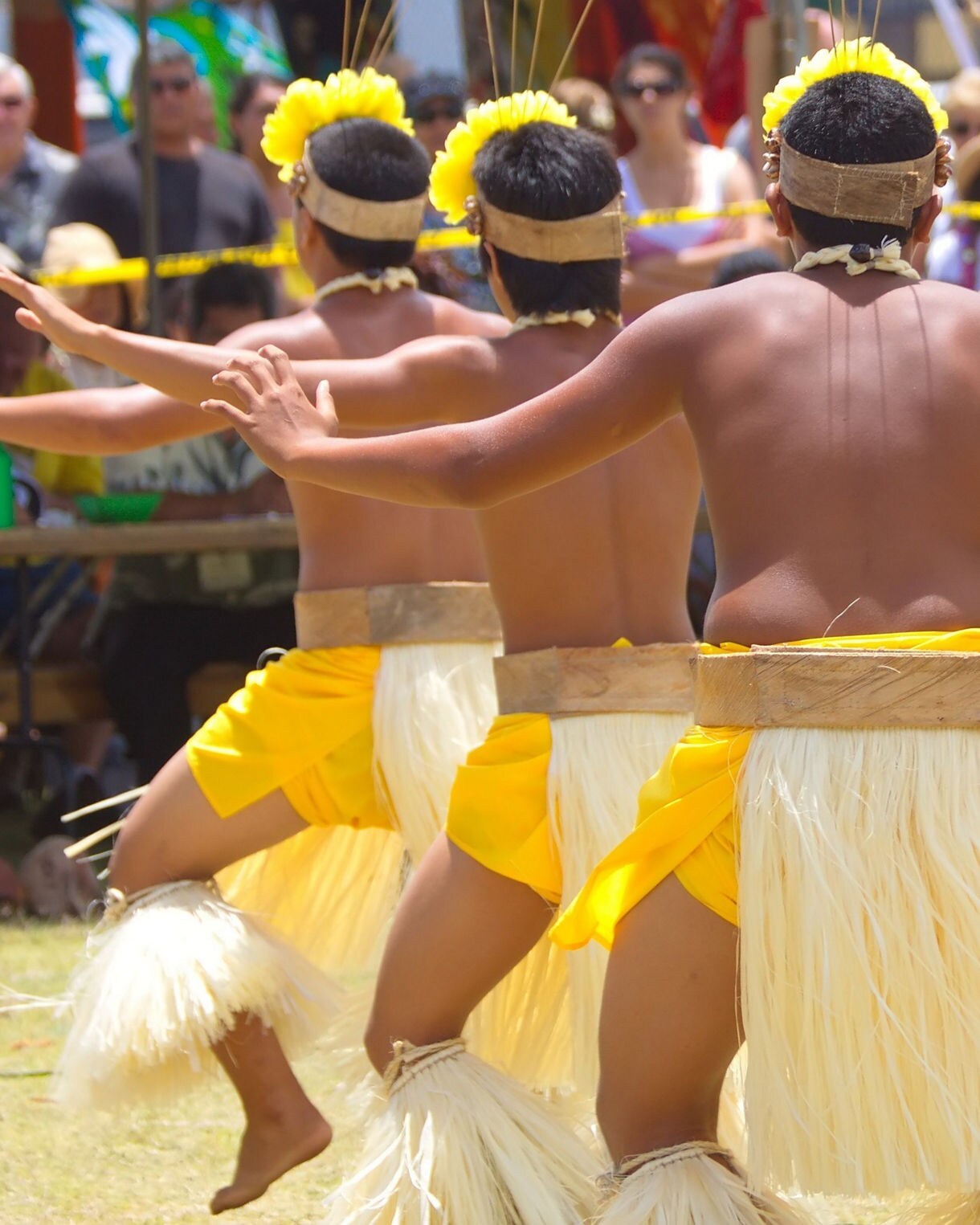 Group of male dancers performing a traditional Hawaiian dance outdoors, wearing yellow skirts and grass adornments before a crowd.