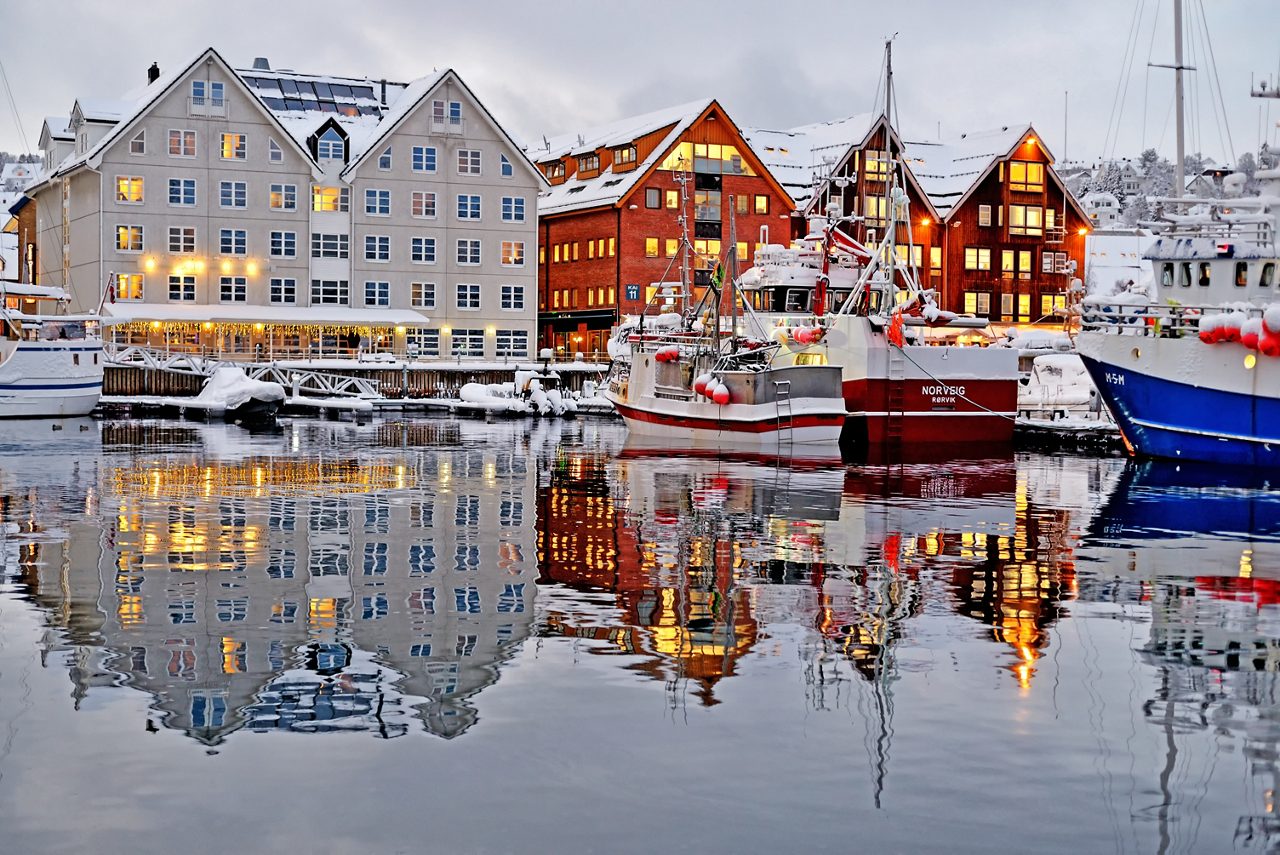 Fishing boats docked at Tromsø port with snowy buildings glowing warmly and reflections on the water.