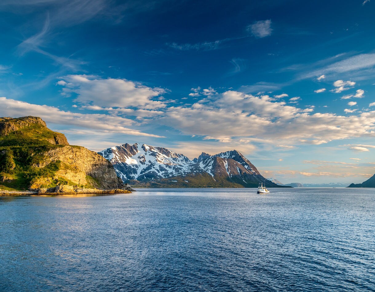 Scenic fjord in Tromsø with a boat on calm blue waters, rugged cliffs and snow-covered mountains in the distance.