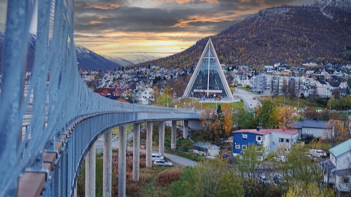 Tromsø Bridge leading to the triangular Arctic Cathedral with mountains and colorful houses at sunset.