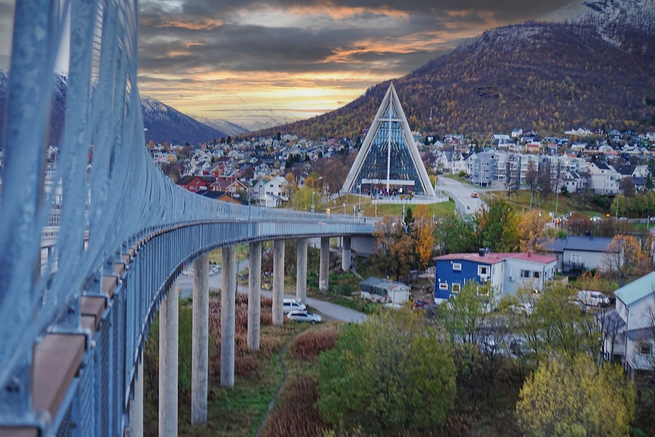 Tromsø Bridge leading to the triangular Arctic Cathedral with mountains and colorful houses at sunset.